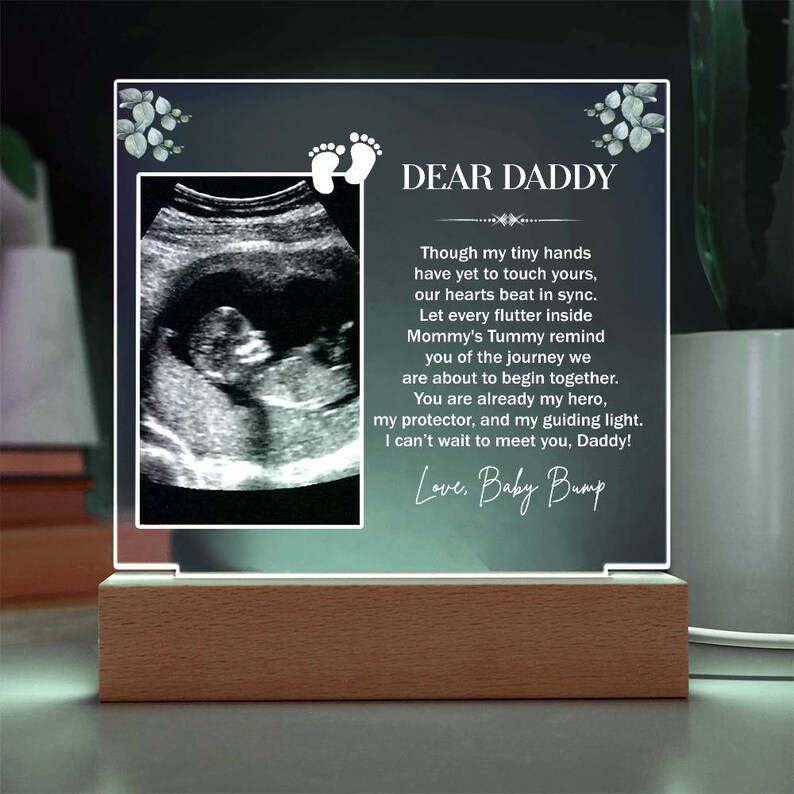 May include: A white acrylic plaque with a black and white ultrasound image and a message to "Dear Daddy" from "Baby Bump". The message reads, "Though my tiny hands have yet to touch yours, our hearts beat in sync. Let every flutter inside Mommy's Tummy remind you of the journey we are about to begin together. You are already my hero, my protector, and my guiding light. I can't wait to meet you, Daddy!"