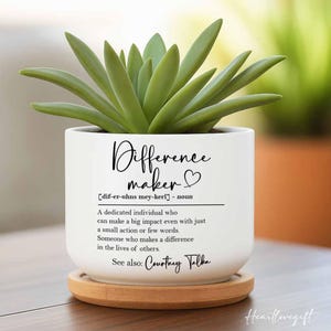 May include: White ceramic planter with a succulent plant. The planter has a black inscription that reads "Difference maker [dif-er-uhns mey-kerl] - noun A dedicated individual who can make a big impact even with just a small action or few words. Someone who makes a difference in the lives of others. See also: Courtney Talka"