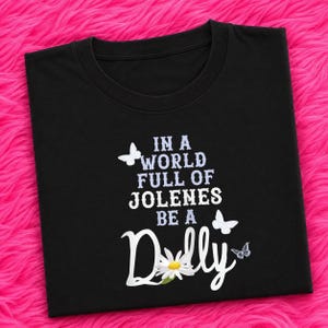 May include: A black t-shirt featuring the white and light purple text IN A WORLD FULL OF JOLENES BE A DOLLY. The design includes three white butterflies and a white daisy flower with a yellow center integrated into the word Dolly.