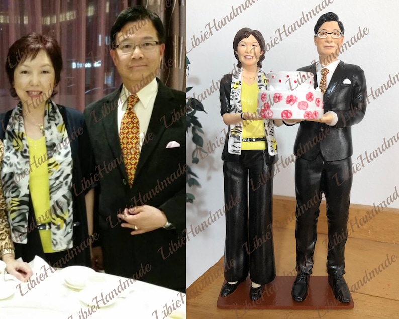 Puede incluir: Una pareja de figuritas personalizadas sosteniendo un pastel blanco con glaseado rosa y flores rojas. La mujer lleva un vestido de estampado animal negro y blanco y una bufanda negra. El hombre lleva un traje negro con camisa blanca y corbata roja.