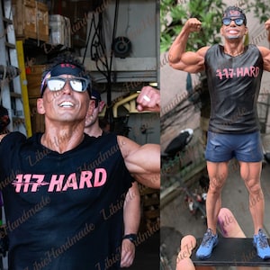 May include: A black sleeveless shirt with the text "117 HARD" printed in pink. The shirt is being worn by a figurine of a muscular man with a black headband and sunglasses. The figurine is standing on a black base.