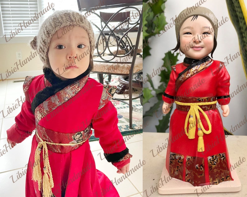 May include: A handmade doll wearing a red and gold Chinese-style robe with a yellow sash and a grey knitted hat. The doll has a realistic face and is standing on a white base.