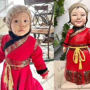 Puede incluir: Una mu&ntilde;eca hecha a mano que lleva una t&uacute;nica roja y dorada de estilo chino con una faja amarilla y un gorro de punto gris. La mu&ntilde;eca tiene una cara realista y est&aacute; de pie sobre una base blanca.
