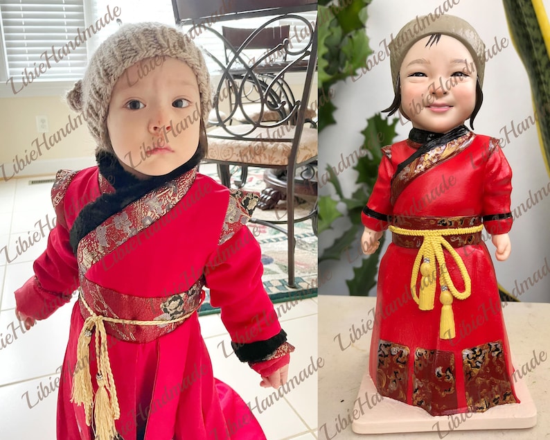 Puede incluir: Una mu&ntilde;eca hecha a mano que lleva una t&uacute;nica roja y dorada de estilo chino con una faja amarilla y un gorro de punto gris. La mu&ntilde;eca tiene una cara realista y est&aacute; de pie sobre una base blanca.