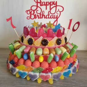 Peut inclure: Un gâteau de bonbons à plusieurs étages décoré de bonbons colorés, notamment des cœurs, des étoiles et des oursons en gélatine. Le gâteau est surmonté d'une bannière rouge "Happy Birthday" et d'une bougie rouge numéro "10".
