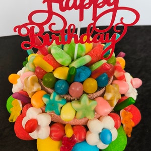 Peut inclure: Un gâteau de bonbons multicolores avec un topper rouge "Joyeux anniversaire". Le gâteau est composé de différents bonbons, dont des oursons en gélatine, des bonbons gélifiés et des guimauves.