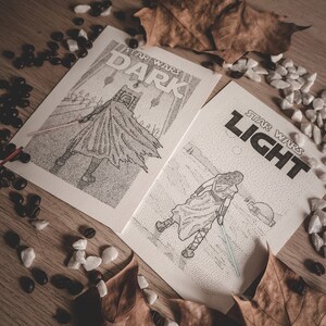 Puede incluir: Dos grabados artísticos de Star Wars en blanco y negro, uno con la etiqueta "DARK" y el otro "LIGHT", que representan personajes con sables de luz. Los grabados están rodeados de hojas de otoño, granos de café y pequeñas piedras blancas sobre una superficie de madera.