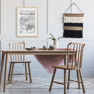 May include: A light wood dining table with a pink tablecloth and two chairs. A framed black and white print of a map with the text "Lord of the Rings" and "Pelennor Fields" is hanging on the wall. A woven wall hanging with black and white fringe is hanging on the wall.