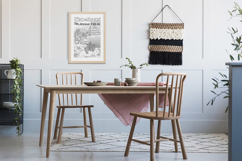 May include: A light wood dining table with a pink tablecloth and two chairs. A framed black and white print of a map with the text "Lord of the Rings" and "Pelennor Fields" is hanging on the wall. A woven wall hanging with black and white fringe is hanging on the wall.