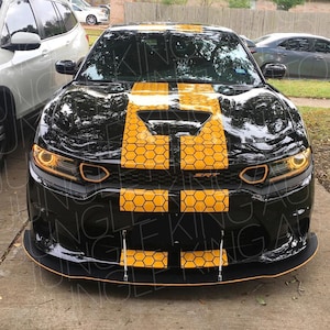 Op de afbeelding: Een zwarte Dodge Charger met een gele en zwarte honingraatstreep decal op de motorkap. De auto heeft een zwarte voorbumper met een zwarte spoiler en een zwarte grille. De auto staat geparkeerd op een verhard oppervlak.