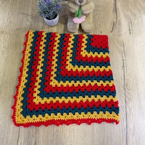 May include: A handmade crochet blanket in a granny square pattern. The blanket features a vibrant color scheme of red, teal, and yellow, with a scalloped red border. A teddy bear and a small potted plant are in the background.