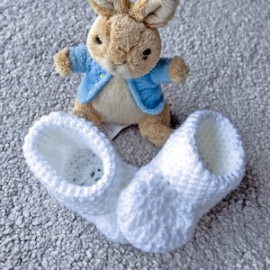May include: A soft toy rabbit in a blue jacket is positioned behind a pair of white, crocheted baby booties. The rabbit has brown fur and long ears. The booties are made of a soft, textured yarn.