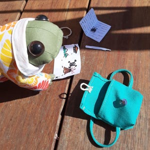 May include: A green frog plush toy wearing a colorful floral print hooded jacket, holding a hand-drawn map with a skull and crossbones, a small white stick, and a turquoise canvas backpack with a zipper.
