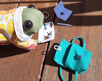 May include: A green frog plush toy wearing a colorful floral print hooded jacket, holding a hand-drawn map with a skull and crossbones, a small white stick, and a turquoise canvas backpack with a zipper.