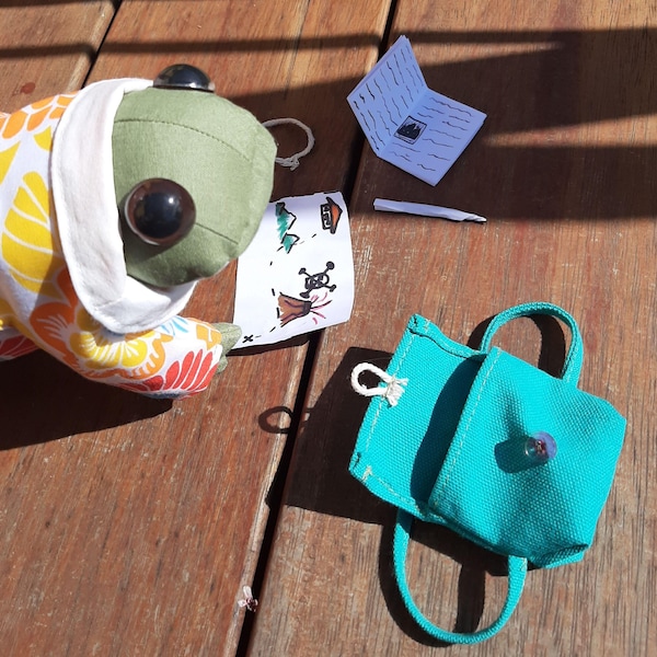 May include: A green frog plush toy wearing a colorful floral print hooded jacket, holding a hand-drawn map with a skull and crossbones, a small white stick, and a turquoise canvas backpack with a zipper.