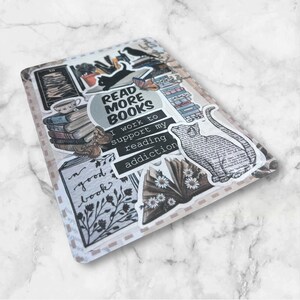 May include: A collage of stickers featuring book-themed illustrations and text. The stickers include a stack of books, a cat, a flower, and text that reads "Read More Books" and "I work to support my reading addiction".