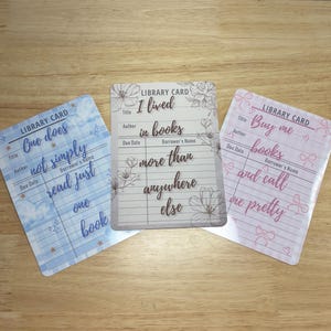 May include: Three decorative library cards with inspirational quotes. One is blue with white text, another is beige with floral accents, and the third is pink with bow graphics. Each card has space for title, author, and borrower's name.