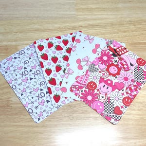 May include: Four different patterned cards with pink and red designs. The designs include strawberries, hearts, bows, smiley faces, and checkered patterns.