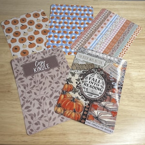 May include: A collection of decorative cards with fall-themed designs. The cards feature patterns of pumpkins, gingham, and floral motifs. One card reads "Cozy Kindle," and another says "Fall Into Reading."