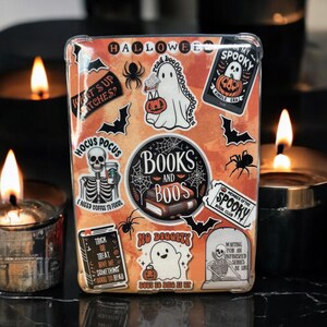 May include: A clear plastic case with a Halloween theme. The case is decorated with colorful stickers featuring ghosts, skeletons, bats, and books. The text on the stickers includes "Hocus Pocus", "Books and Boos", "No Diggity", and "My Spooky Book Club".