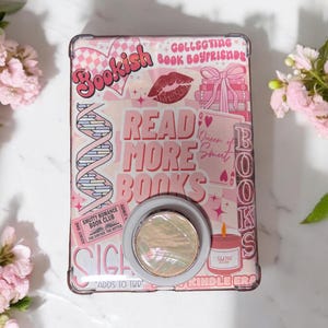 Może przedstawiać: Różowa i biała obudowa Kindle z uchwytem. Obudowa zawiera napisy "Bookish", "Read More Books" i inne grafiki. Do tyłu obudowy przymocowany jest okrągły, perłowy uchwyt. Obudowa jest ozdobiona sercami, ustami i motywami związanymi z książkami.