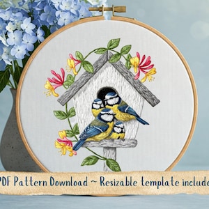 Puede incluir: Diseño bordado de una casita de pájaros con tres pájaros azules posados en el borde. La casita de pájaros es blanca con un techo gris y está rodeada de enredaderas verdes y flores rosas y amarillas.
