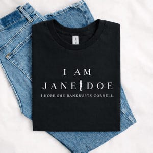 May include: A black short-sleeved t-shirt featuring white text that reads I AM JANE DOE with a small silhouette of a person replacing the letter I in Jane, and the additional text I HOPE SHE BANKRUPTS CORNELL printed below in white.
