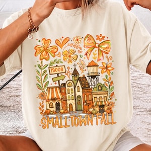 Pode incluir: Uma t-shirt de cor creme com uma impressão gráfica colorida de uma cena de cidade pequena com edifícios, uma torre de água, flores e um laço. O texto "19008" aparece numa placa e as palavras "SMALL TOWN FALL" estão impressas na parte inferior.