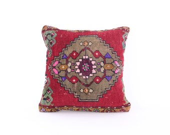 May include: Square decorative pillow with a red background and a central geometric design in shades of olive green, purple, pink, and gold. The pillow has a traditional rug pattern.
