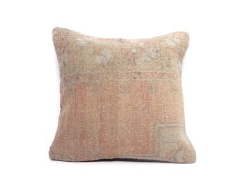 May include: A square cushion with a faded, vintage aesthetic. The cushion displays a mix of muted colours, including beige, pink, and touches of blue. The design has a patchwork or distressed appearance, possibly handmade or antique.