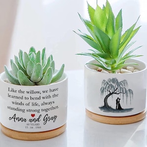 Puede incluir: Dos macetas de cerámica blanca con bases de madera, cada una con una suculenta. Una maceta tiene el texto "Anna and Gray" y una fecha. La otra presenta una silueta de una pareja bajo un árbol. Ambas macetas están llenas de pequeñas piedras blancas.