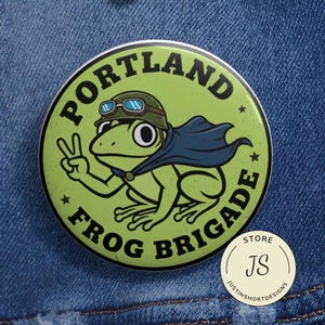May include: A round, green button with the words "PORTLAND FROG BRIGADE" encircling a cartoon frog wearing a helmet and cape, giving a peace sign. The button is on a denim background. A smaller, round button with "STORE JS" is also visible.