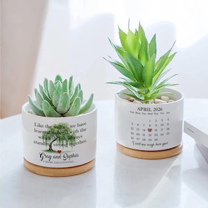 May include: Two white ceramic planters with succulents, each on a wooden base. One features text about bending with life's winds, the other a calendar for April 2026. The planters are filled with small white stones.