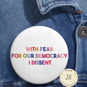 Può includere: Un bottone rotondo bianco con la scritta "WITH FEAR FOR OUR DEMOCRACY I DISSENT" in vari colori. Il bottone è posto su uno sfondo di jeans blu. Un piccolo logo del negozio si trova nell'angolo in basso a destra.