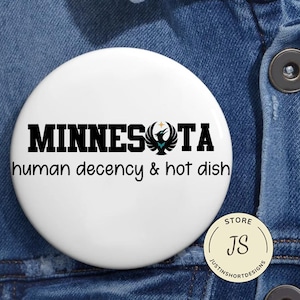 Può includere: Un bottone rotondo bianco con la parola "MINNESOTA" in grassetto nero, una grafica di uccello nero e verde acqua e la frase "human decency & hot dish" in corsivo nero. Un piccolo bottone color oro con "STORE" e "JS" si trova in basso a destra.