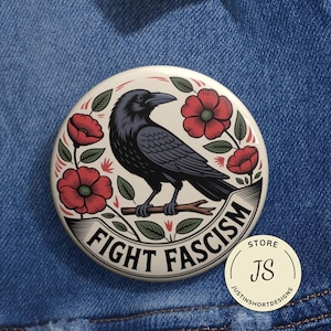 Puede incluir: Un botón redondo con una ilustración de un cuervo negro, flores rojas y la frase "FIGHT FASCISM" en una pancarta. El botón está sobre un fondo de mezclilla azul. Un pequeño logotipo de la tienda está en la esquina inferior derecha.