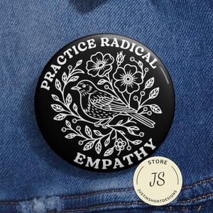 Może przedstawiać: Czarny guzik z białym napisem "PRACTICE RADICAL EMPATHY" wokół wzoru ptaka i kwiatów. Mniejsza, okrągła, beżowa naklejka z "JS" i "justinshortdesigns" znajduje się w prawym dolnym rogu.