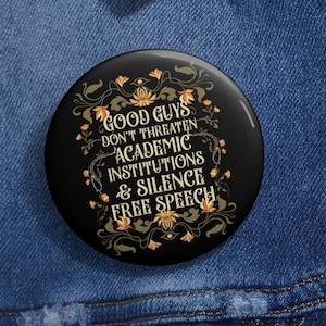 Pode incluir: Um botão redondo preto com o texto "GOOD GUYS DON'T THREATEN ACADEMIC INSTITUTIONS & SILENCE FREE SPEECH" em letras brancas e douradas. O botão tem um design floral e está sobre um fundo de ganga azul.