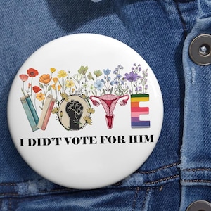 May include: White button with a colorful floral design, a raised fist, a uterus, and the word "VOTE" spelled out with rainbow stripes. The text "I didn't vote for him" is printed below the design.