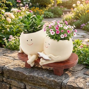 May include: Two cream-colored ceramic planters with smiling faces, one holding a book. One planter holds a green succulent, the other has pink and white flowers. They sit on a brown bench, with a garden background.