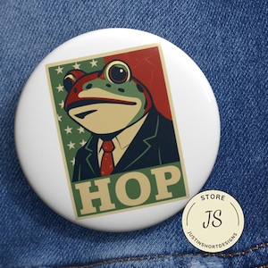 Puede incluir: Botón blanco con una ilustración de rana con traje, la palabra "HOP" en letras verdes y un fondo rojo y verde. La rana tiene ojos grandes y una corbata roja. Un pequeño logotipo de la tienda está en la esquina inferior derecha.