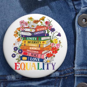 May include: A white button featuring a rainbow-coloured word "EQUALITY" at the bottom. The word is surrounded by a stack of colourful books with words such as "Justice", "Compassion", "Unity", "Diversity", "Change", "Kindness", "Inclusion", "Peace", and "Love" printed on them. The books are surrounded by colourful flowers.