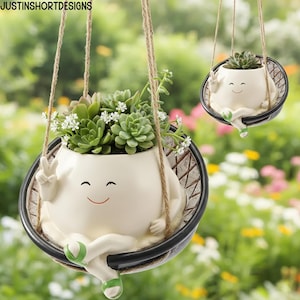 Puede incluir: Dos macetas colgantes de cerámica con un diseño de cara sonriente, cada una con suculentas y pequeñas flores blancas. Las macetas están suspendidas por una cuerda y tienen un borde negro con un patrón tejido. Las macetas son de color crema con detalles verdes.