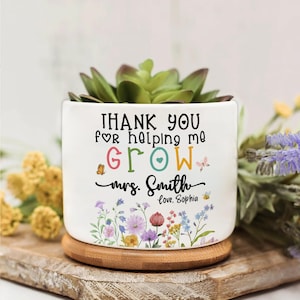 Puede incluir: Maceta de cerámica blanca con una suculenta, con el texto "THANK YOU for helping me GROW Mrs. Smith Love, Sophia" y diseños florales. Descansa sobre un posavasos de madera.