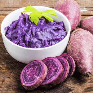 May include: A white bowl filled with vibrant purple mashed sweet potatoes, garnished with a sprig of green celery. Sliced purple sweet potatoes and whole sweet potatoes are arranged on a wooden surface, showcasing the ingredients.