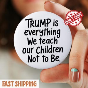 Puede incluir: Un botón blanco con el texto "TRUMP is everything We teach our Children Not to Be." en negro. Es visible un sello rojo "DON'T COPY". El botón lo sostiene una persona.