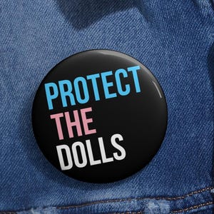 Può includere: Un bottone nero con il testo "PROTECT THE DOLLS" in lettere blu, rosa e bianche.