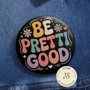 Puede incluir: Un botón redondo negro con las palabras "BE PRETTI GOOD" en letras coloridas de estilo retro. El botón presenta detalles florales y de corazón. Un pequeño logotipo circular de la tienda está en la parte inferior.