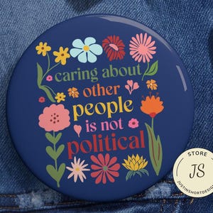May include: A navy blue button with the text "caring about other people is not political" surrounded by colorful flowers. The button has a glossy finish and is set against a denim background.