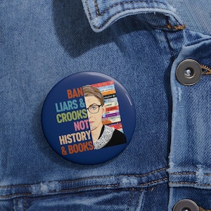 May include: A blue button with a portrait of Ruth Bader Ginsburg and the text "Ban Liars & Crooks Not History & Books".